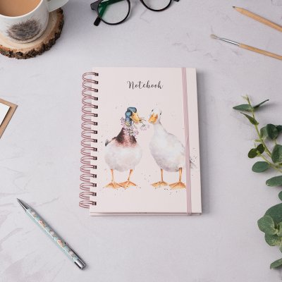 A pair of ducks carrying daisies on an A5 spiral bound notebook on desk surrounded by stationery and a hot drink 